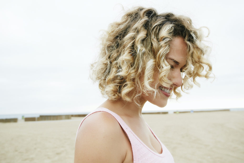 Portrait of smiling young woman with blond curly hair standing on sandy beach.