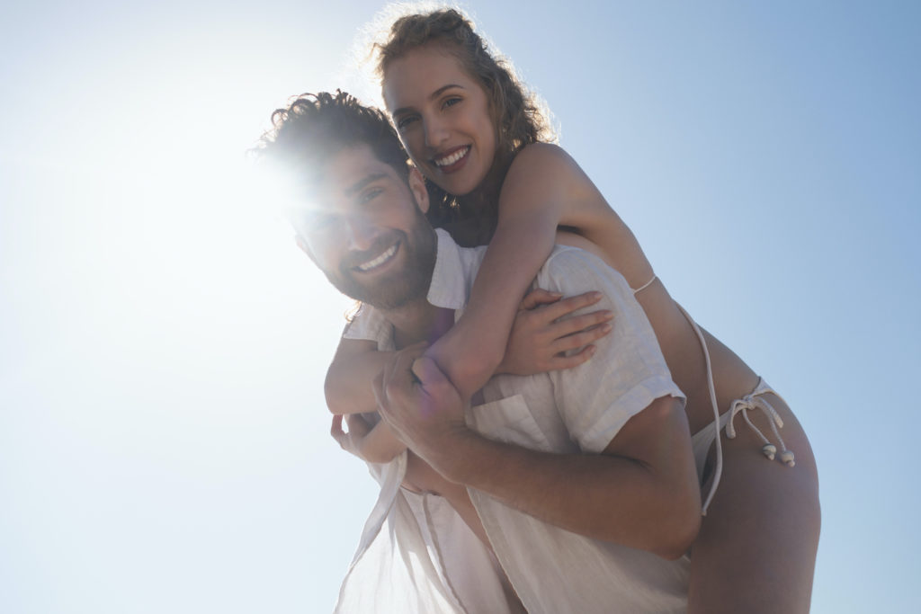 Portrait of young Caucasian man carrying pretty Caucasian woman piggyback at beach. They are smiling and looking at camera