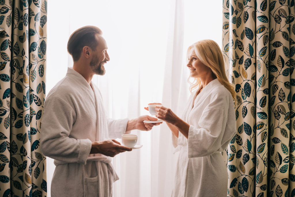 side view of happy middle aged couple in bathrobes drinking coffee and smiling each other in hotel room